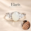 Elaris Moonstone Ring featuring an oval white moonstone center stone with diamond accents on a silver band, shown on a warm beige textured background with a small inset of the ring worn on a hand.